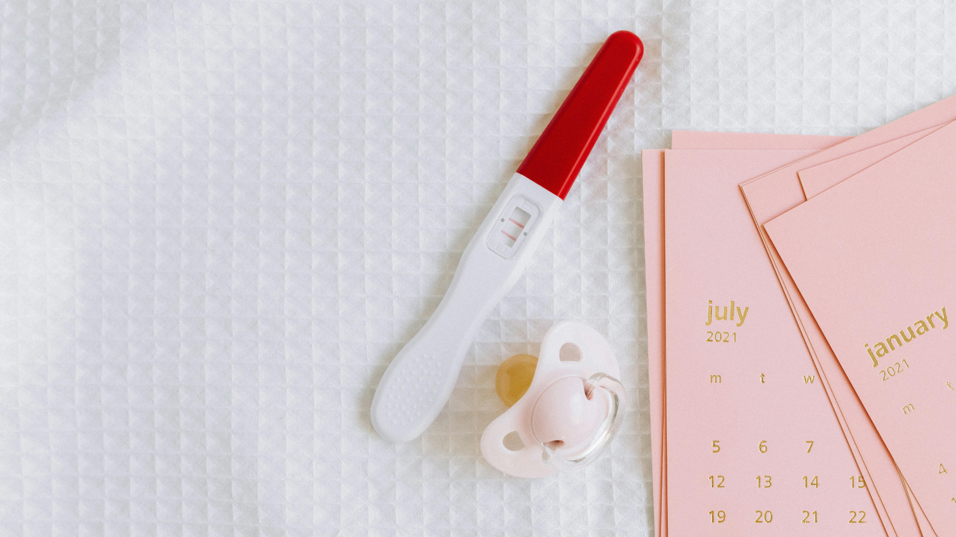 A positive pregnancy test with a red cap, a pink pacifier, and pastel pink calendars displaying months from 2021 are arranged on a textured white fabric surface.