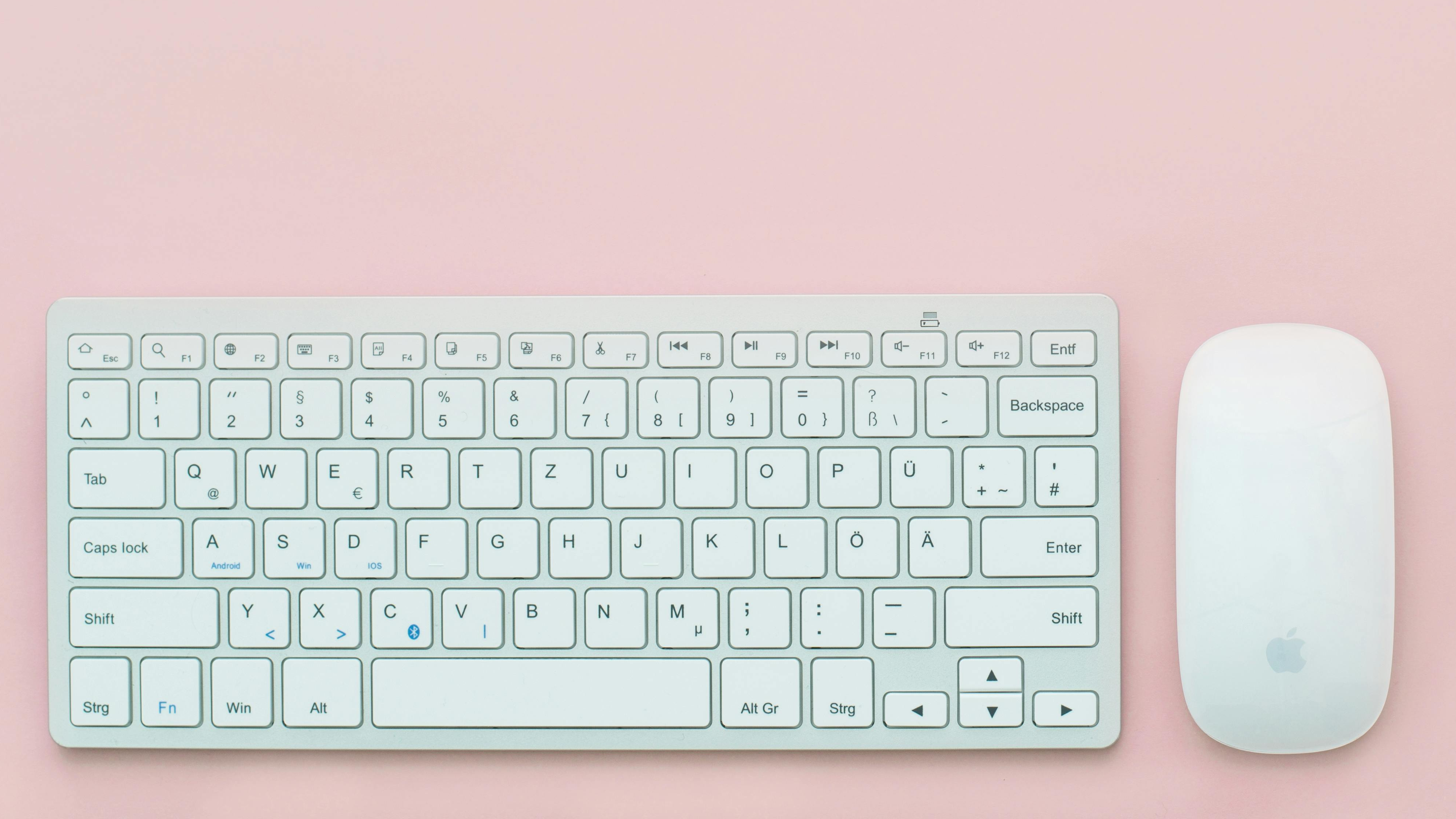 A white wireless keyboard with a German QWERTZ layout and an Apple Magic Mouse are placed on a pastel pink background in a minimalistic flat-lay arrangement.